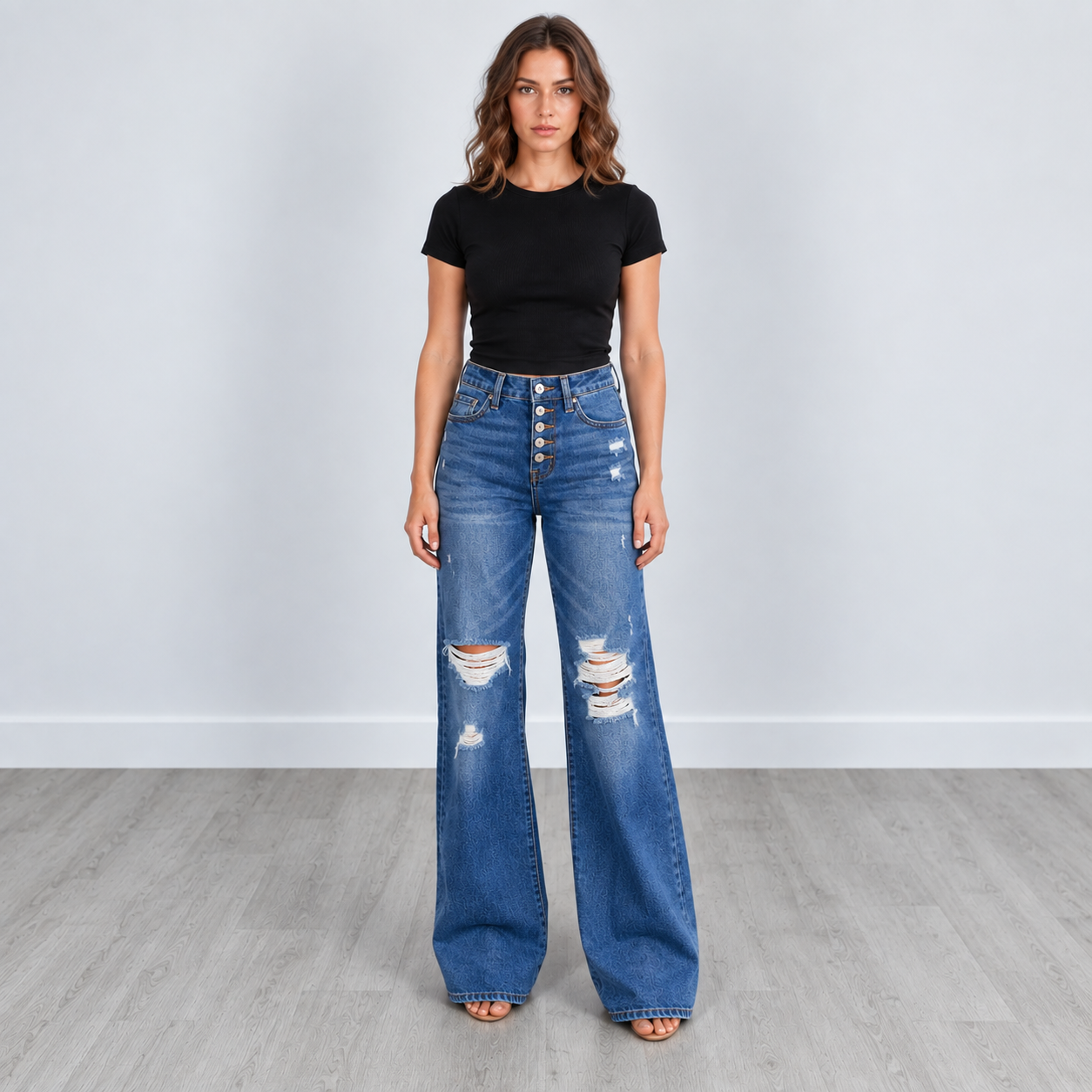 A woman stands in a studio wearing high-waisted, wide-leg blue denim jeans with distressed rips and button fly details, paired with a black short-sleeve crop top.