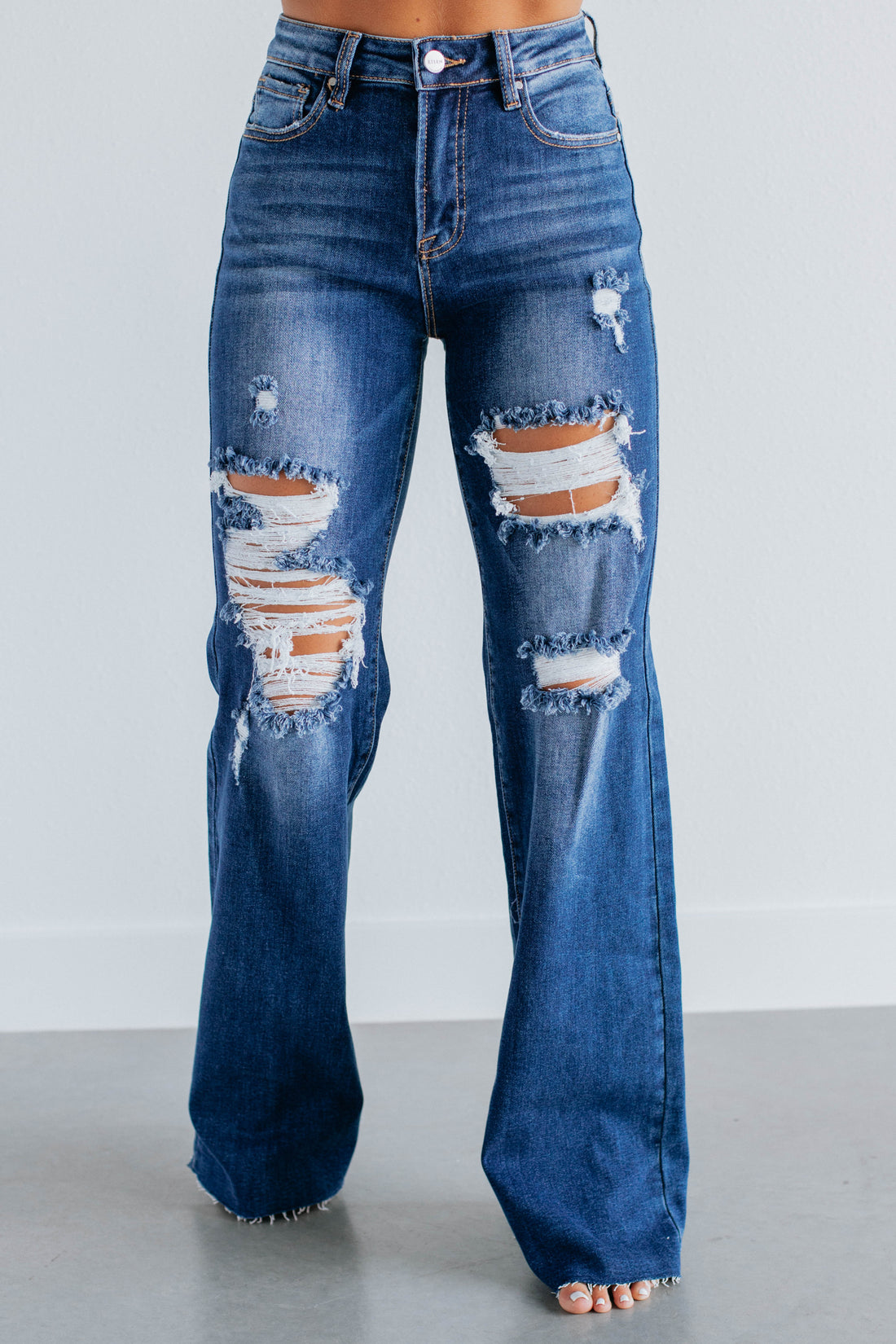 A person wearing high-waisted, wide-leg blue denim jeans with heavy distressing and ripped details on the thighs and knees, shown from the waist down against a light gray studio background.