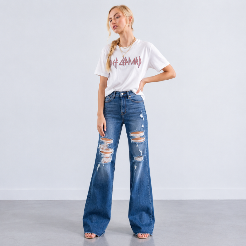 A woman modeling a white graphic t-shirt and distressed wide-leg blue jeans with ripped details on the thighs, standing against a light gray studio background.