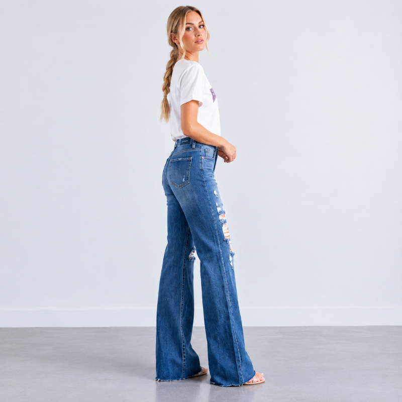 A woman stands in profile wearing blue high-waisted flare jeans with distressed rips on the front, paired with a white t-shirt, against a light gray studio background.
